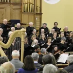 Group of singers in front of a pipe organ singing accompanied by a small stringed orchestra